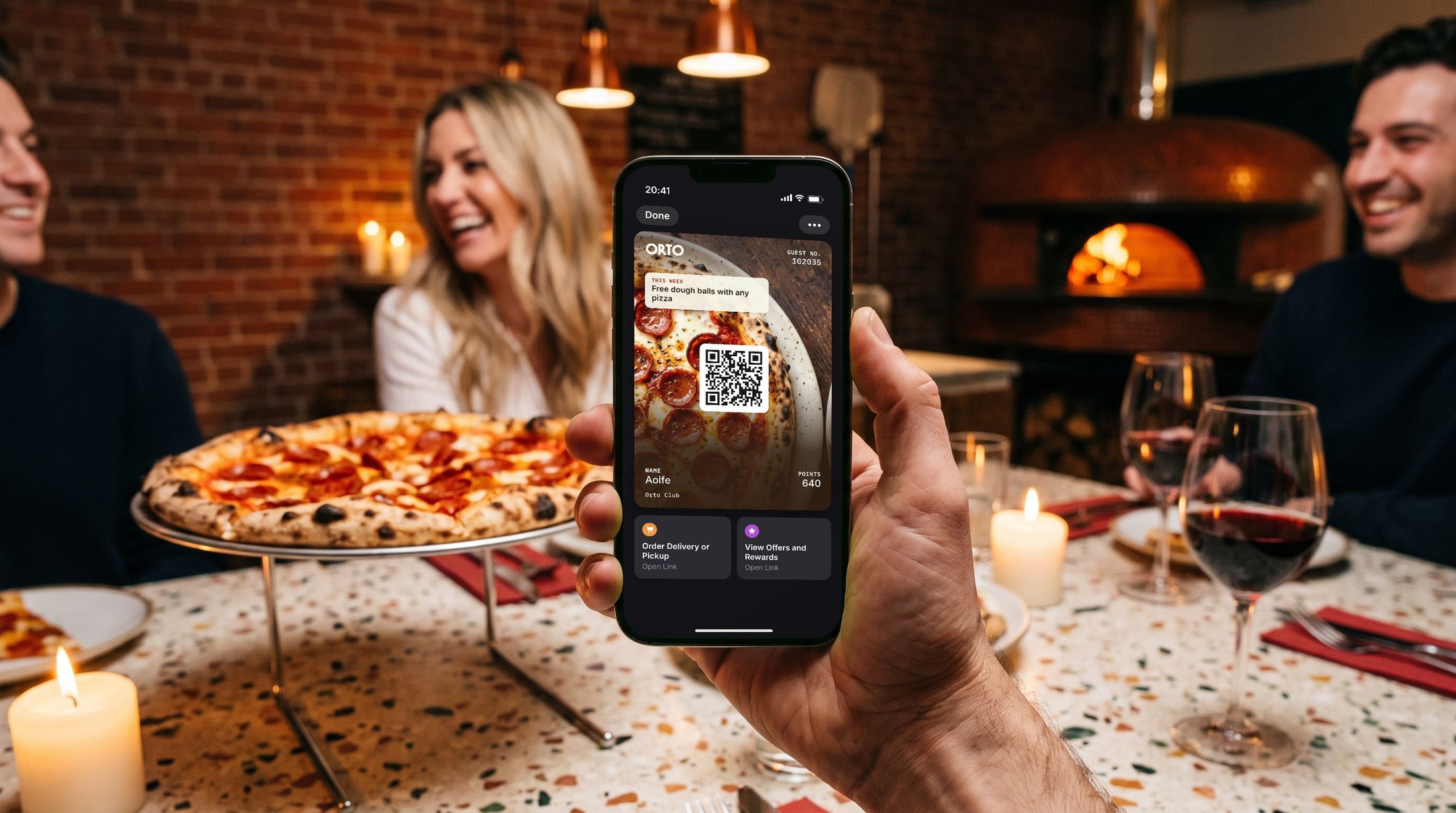 A guest at the table holds up a phone showing the Orto Club wallet pass